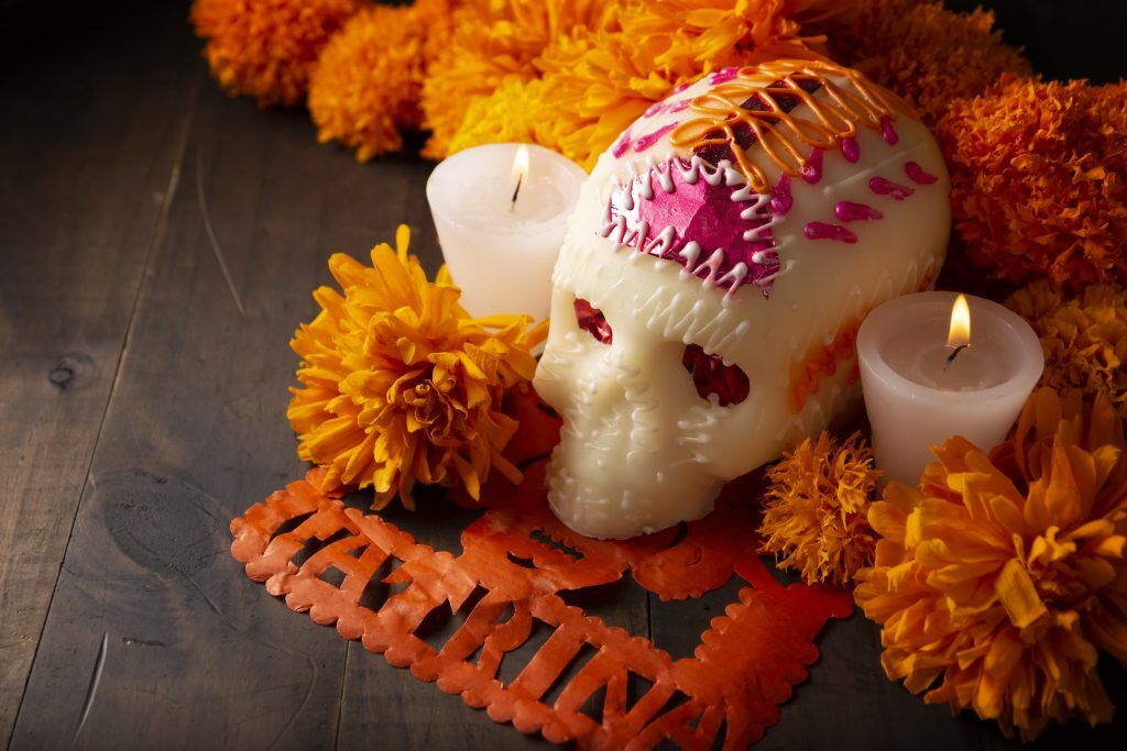 A Calavera Sugar Skull with red and orange adornment surrounded by candles and orange flowers.